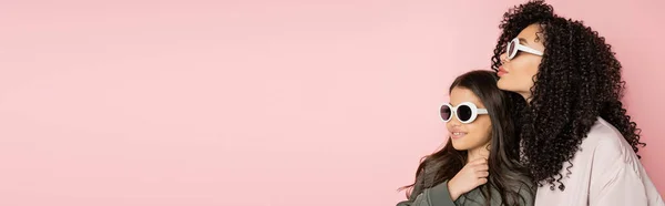 Donna riccia in piedi vicino a figlia preadolescente in occhiali da sole isolati su rosa, banner — Foto stock