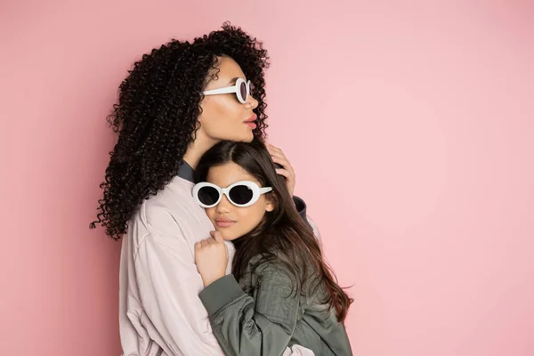 Elegante donna abbracciando figlia in occhiali da sole su sfondo rosa — Foto stock