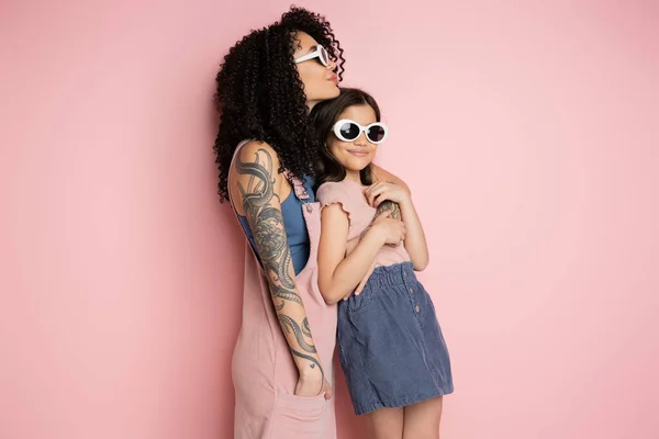 Tattooed young woman hugging daughter in sunglasses on pink background - foto de stock