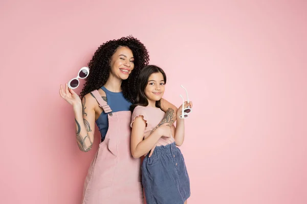 Tattooed woman hugging daughter and holding sunglasses on pink background - foto de stock
