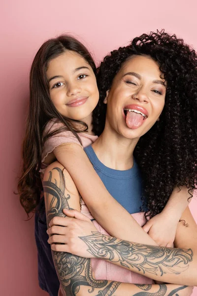 Lächelndes Mädchen umarmt tätowierte Mutter, die ihre Zunge auf rosa Hintergrund ausstreckt — Stockfoto