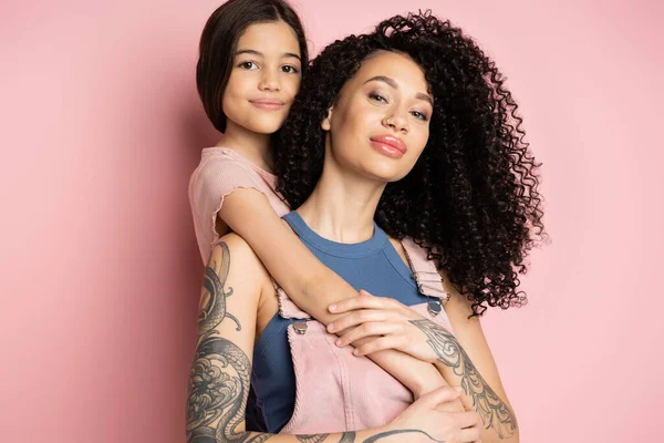 Sonriente niño abrazando rizado y tatuado madre sobre fondo rosa - foto de stock