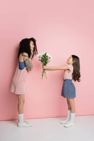 Side view of smiling girl giving flowers to trendy mom on pink background — Stock Photo
