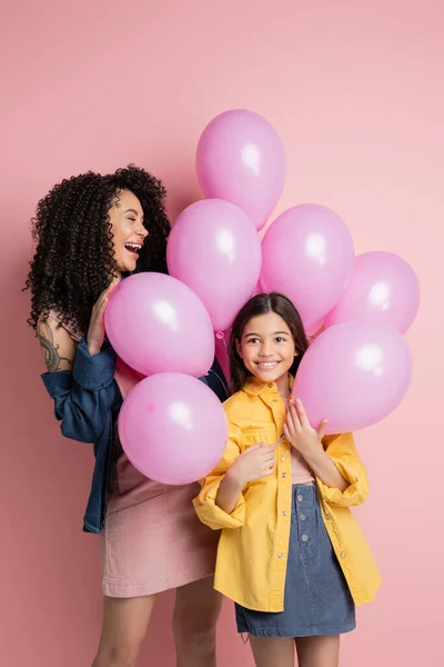 Positive mother and stylish kid standing near festive balloons on pink background - foto de stock