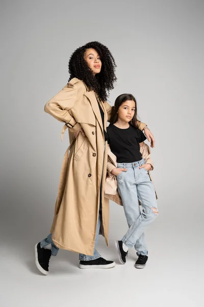 Full length of stylish mother and daughter in trench coats posing on grey background — Stock Photo