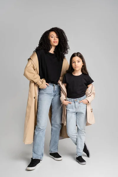 Lockige Mutter im Trenchcoat posiert neben stilvoller Tochter auf grauem Hintergrund — Stockfoto