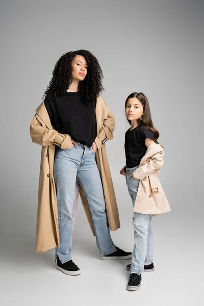 Preteen child in trench coat posing near stylish mom on grey background — стоковое фото
