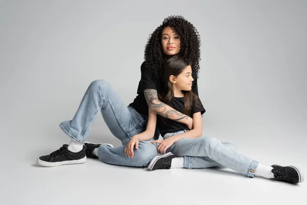 Stylish mother hugging daughter in jeans and t-shirt on grey background — стоковое фото