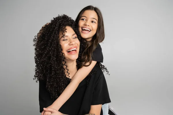 Cheerful girl hugging curly mom in black t-shirt isolated on grey — Stock Photo
