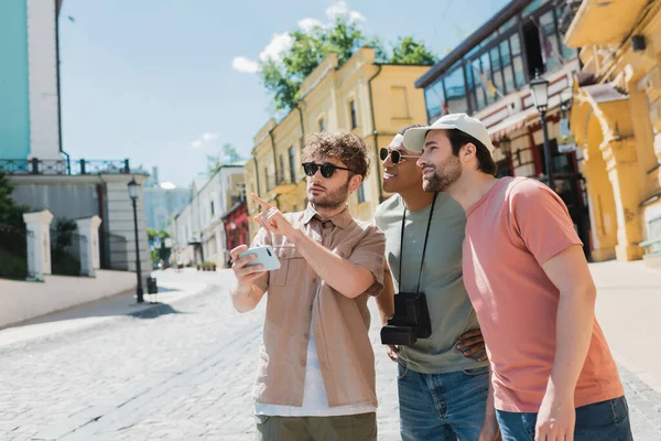 Young tour guide holding mobile phone and pointing with finger near interracial tourists on Andrews descent in Kyiv — Stock Photo