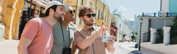 Giovane guida in occhiali da sole in possesso di smartphone e puntando con il dito vicino ai turisti multietnici sulla discesa Andrews a Kiev, banner — Foto stock