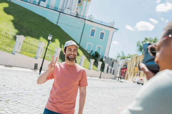 Homme afro-américain flou avec caméra vintage prenant une photo de touriste barbu heureux montrant signe de victoire sur la descente Andrews à Kiev — Photo de stock