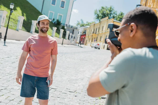 Размытый африканский американец с винтажной камерой фотографирует улыбающегося друга на спуске Эндрюса в Киеве — стоковое фото