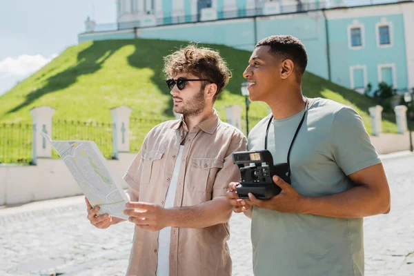 Guida turistica in occhiali da sole guardando la mappa vicino al viaggiatore afroamericano con fotocamera vintage sulla discesa Andrews a Kiev — Foto stock