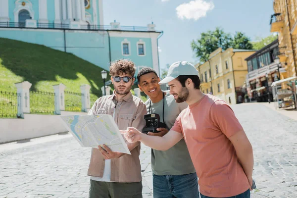 Turista barbudo apuntando al mapa de la ciudad cerca de guía turístico y hombre afroamericano sonriente con cámara vintage en el descenso de Andrews en Kiev - foto de stock