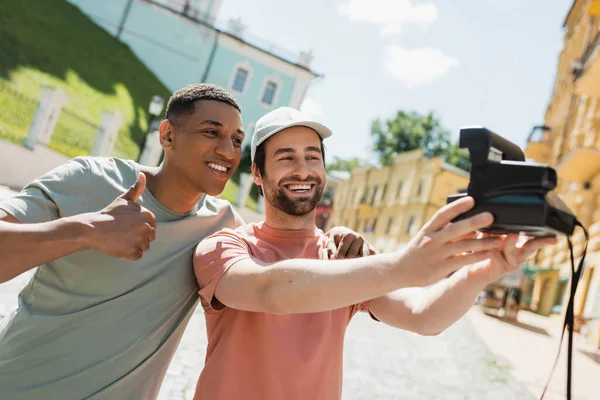 Happy african american man showing thumb up while friend taking selfie on vintage camera on Andrews descent in Kyiv — Stock Photo