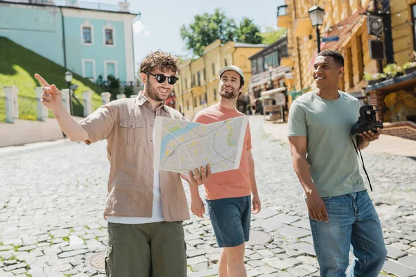 Smiling guide in sunglasses looking at map and pointing with finger near multiethnic tourists walking on Andrews descent in Kyiv — Stock Photo