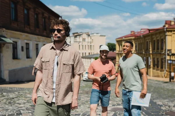 Tour guide in sunglasses and headset walking near blurred multicultural tourists on Andrews descent in Kyiv — Stock Photo