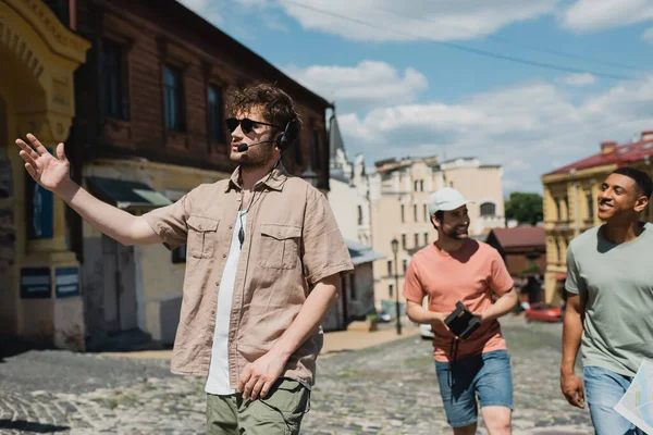 Giovane guida in auricolare e occhiali da sole che punta con mano durante l'escursione con uomini interrazziali sul quartiere Podil a Kiev — Foto stock