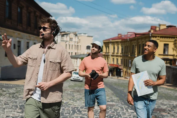 Lächelnde multikulturelle Touristen mit Reisekarte und Oldtimer-Kamera, die mit Führer im Headset auf dem historischen Viertel in Kiew spazieren gehen — Stockfoto