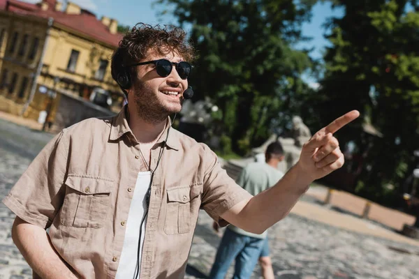 Guia turístico alegre em headset e óculos de sol apontando com o dedo na descida Andrews borrada em Kiev — Fotografia de Stock