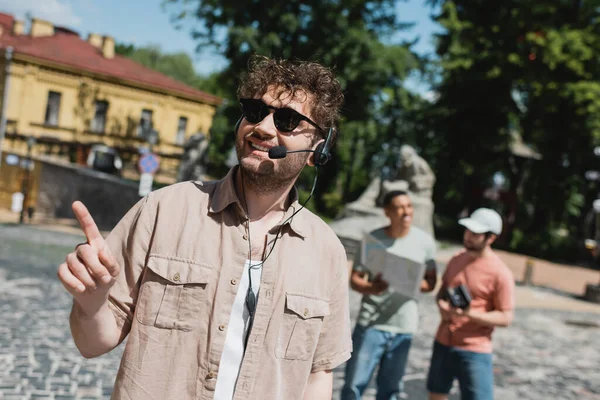 Guia jovem e despreocupado em óculos de sol e fone de ouvido apontando com o dedo perto turvo turistas inter-raciais no distrito de Podil, em Kiev — Fotografia de Stock