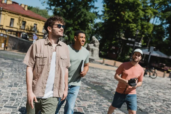 Turistas multiétnicos sonrientes con cámara vintage y mapa caminando con guía turístico en el descenso de Andrews en Kiev - foto de stock