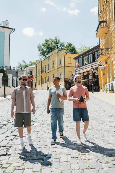 Multiethnische Touristen mit Karte und Oldtimer-Kamera gehen mit Reiseleiter auf dem Bürgersteig der Andreasabfahrt in Kiew — Stockfoto