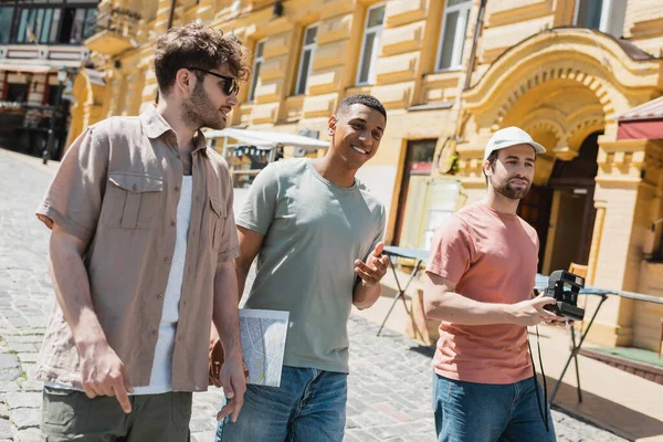 Joyful interracial travelers with city map and vintage camera walking with tour guide in sunglasses on Andrews descent in Kyiv — Stock Photo