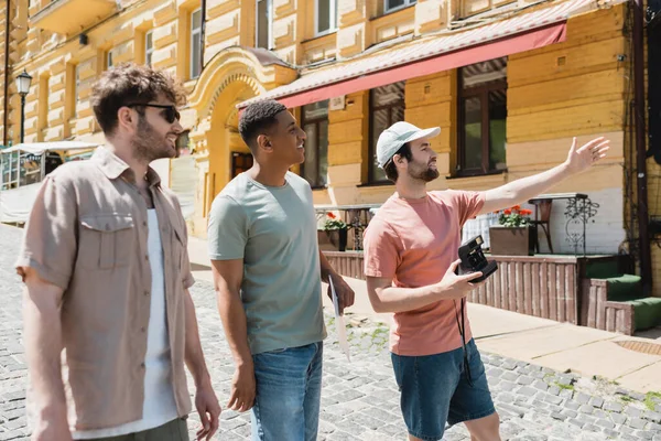 Barbuto turista in berretto solare tenendo fotocamera vintage e puntando con mano vicino amici interrazziale sulla discesa Andrews a Kiev — Foto stock