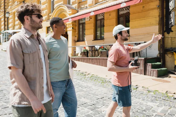 Side view of tourist with vintage camera pointing with hand during excursion with interracial friends on Andrews descent in Kyiv — Stock Photo