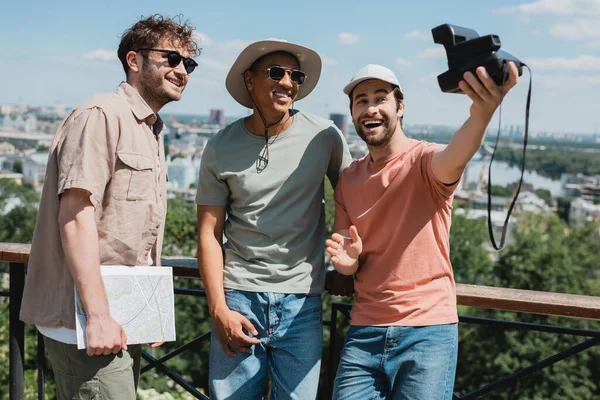 Carefree bearded man with vintage camera taking selfie with african american friend and tour guide near fence in city park — Stock Photo