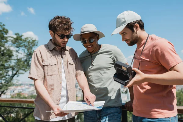 Young tour guide in sunglasses looking at travel map near interracial tourists with vintage camera in city park — Stock Photo