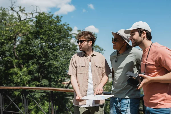 Happy multiethnic travelers in sun hats looking away near tour guide with map in urban park — Stock Photo