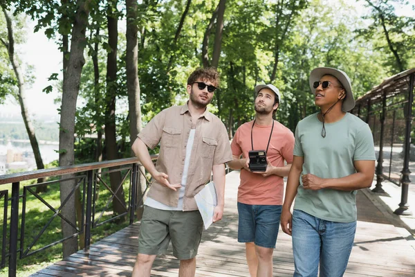 Multicultural travelers in sun hats looking away during summer walk with tour guide in urban park — Stock Photo