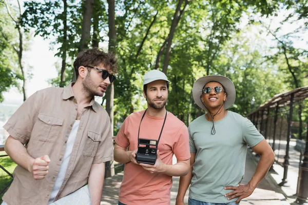 Молодий екскурсовод в сонцезахисних окулярах розмовляє з багатоетнічними туристами в сонячних капелюхах під час екскурсії в міському парку — стокове фото