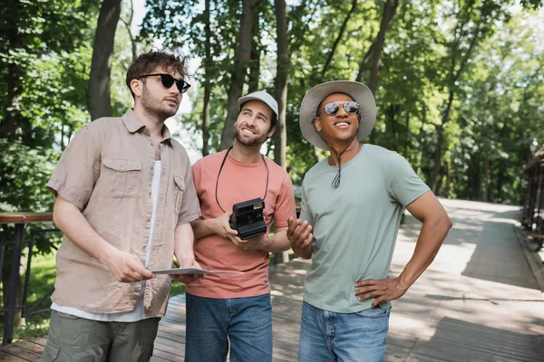 Веселий бородатий турист з старовинною камерою, що дивиться далеко від мультикультурних чоловіків у зеленому міському парку — стокове фото