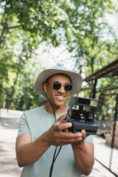 Smiling african american traveler in sun hat and sunglasses taking photo on vintage camera in summer park — Stock Photo