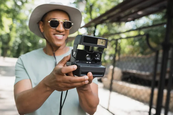 Enfoque selectivo de la cámara vintage en manos del turista afroamericano feliz en gafas de sol tomando fotos en el parque - foto de stock