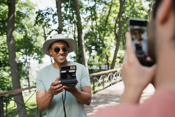 Blurred man with smartphone taking photo of carefree african american tourist in sunglasses posing with vintage camera in park — Stock Photo