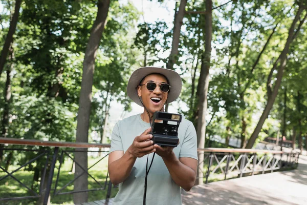 Stupéfait voyageur afro-américain portant des lunettes de soleil et chapeau de soleil tout en prenant des photos sur la caméra vintage sur la passerelle dans le parc — Photo de stock