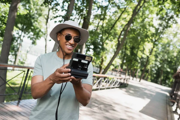 Heureux touriste afro-américain en chapeau de soleil et lunettes de soleil prenant des photos sur caméra vintage sur passerelle dans le parc de la ville — Photo de stock