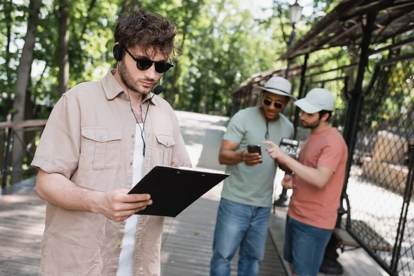 Tour guide in sunglasses and headset looking at route on clipboard near blurred interracial tourists wearing sun hats in city park — Stock Photo