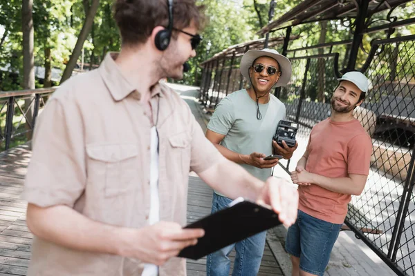 Hombres despreocupados multiétnicos con cámara vintage y teléfono inteligente mirando borrosa guía turístico con portapapeles en el parque de verano - foto de stock