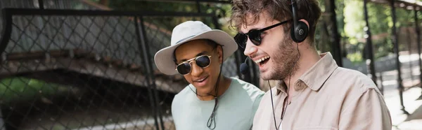 Молодий гід в сонцезахисних окулярах і гарнітурі посміхається біля афроамериканського туриста в сонячному капелюсі, банер — стокове фото