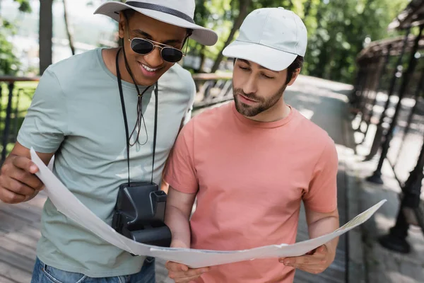 Улыбающийся африканский американский путешественник с винтажной камерой смотрит на карту путешествий с другом в летнем парке — стоковое фото