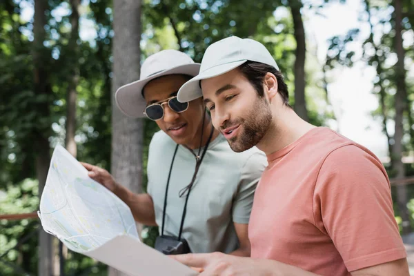 Cheerful multiethnic tourists in sun hats looking at travel map in blurred park — Stock Photo