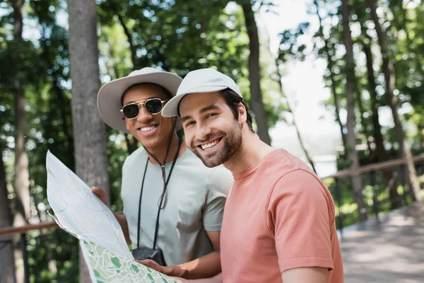 Turistas multiétnicos despreocupados en sombreros con mapa de viaje y mirando a la cámara en el parque de verano - foto de stock