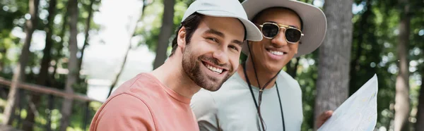 Fröhliche Touristin blickt in die Kamera neben einem afrikanisch-amerikanischen Freund mit Sonnenbrille und hält Reisekarte im Park, Banner — Stockfoto