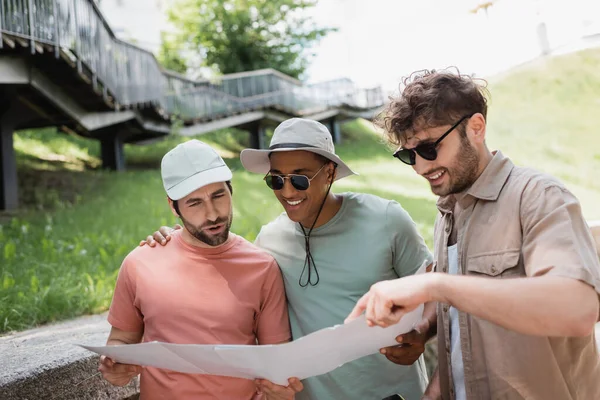 Cheerful man in sunglasses pointing at map near interracial friends in sun hats on urban street — Stock Photo